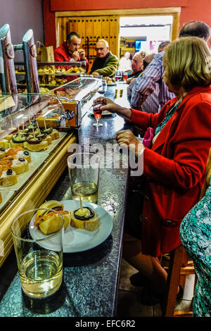 Tapas bar interno in Spagna. I clienti si trovano al banco dove mangiano pinchos e bevono vino o birra. Vetrina in vetro con ampia scelta di tapas visibili Foto Stock