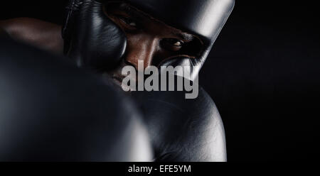 Close-up di immagine africana boxer maschio indossa testa protettiva guard e guanti di protezione contro lo sfondo nero. Giovane uomo che esercitano box Foto Stock