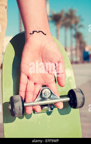 Primo piano di un giovane uomo con i baffi tatuati nel suo polso tenendo uno skateboard Foto Stock