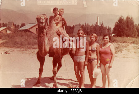 Il 12 Dic. 2014 - Kirghizistan - circa 1983: Riproduzione di un antico mostra fotografica di uomo e di due donne in posa vicino a un cammello, su cui sedersi due ragazze contro lo sfondo delle montagne e la ricreazione. Iscrizione in russo: Issyk-Kul Lago - il lago più grande in Kirghizistan, undrained, parte dei 25 laghi più grandi del mondo per area e 7 nell'elenco dei più profondi laghi. (Credito Immagine: © Igor Golovniov/ZUMA filo/ZUMAPRESS.com) Foto Stock