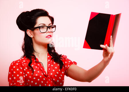 Rétro. Donna elegante studente o insegnante in occhiali lettura libro su rosa. Bruna ragazza in stile pinup. L'istruzione. Foto Stock