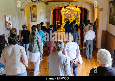 Hare Krishna devoti stand a pregare e celebrare nella parte anteriore di un altare in un tempio di camera. Foto Stock