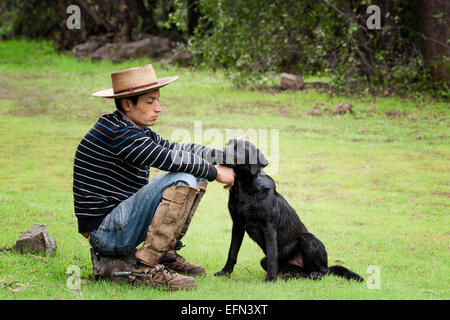 Cowboy cileno (arriero) animali domestici il suo cane nero come si siede su una roccia nella valle di El Toyo regione del Cajon del Maipo, Cile, Sud un Foto Stock