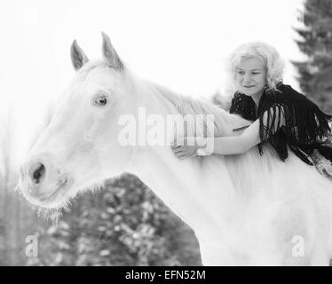Attraente donna bionda che abbracci un cavallo bianco, coperto giornata invernale Foto Stock