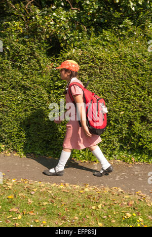 Giovane ragazza in rosso la scuola primaria uniforme di camminare lungo il sentiero accanto a verde e lussureggiante privet hedge indossando hat per tenere fuori il sole Foto Stock