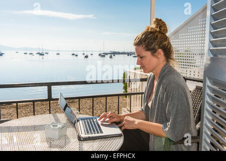 Donna al lavoro su il suo computer portatile su un balcone che si affaccia sull'oceano, Port de Pollenca, Maiorca, isole Baleari, Spagna Foto Stock