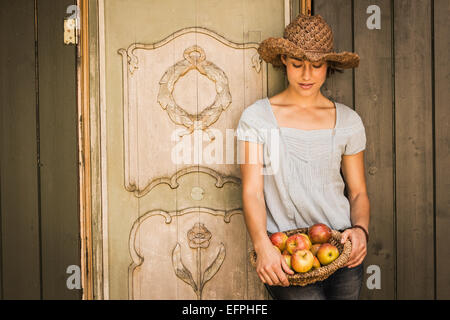 Giovane donna che indossa cappello di paglia, appoggiata contro un capannone, cestello di contenimento di mele Foto Stock
