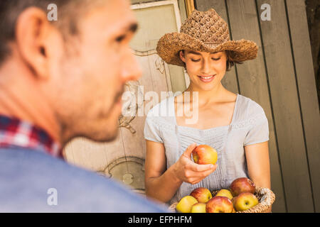 Giovane donna che indossa cappello di paglia, azienda apple dal cestello, metà uomo adulto in foregound Foto Stock