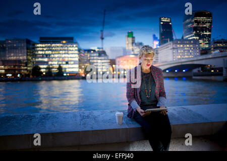 Coppia imprenditrice seduta sul Tamigi waterfront utilizzando tavoletta digitale di notte, Londra, Regno Unito Foto Stock