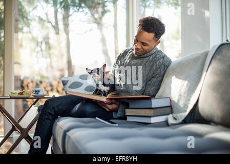 Uomo che indossa un rullo grigio-collo ponticello seduta su un divano con un cane sul suo giro guardando un libro. Foto Stock