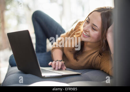 Donna sdraiata su un divano guardando il suo laptop, sorridente. Foto Stock