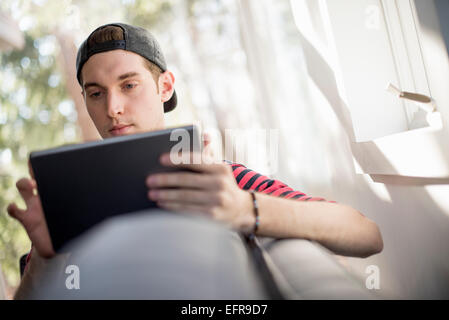 Uomo che indossa un cappello da baseball indietro, seduto su un divano, guardando una tavoletta digitale. Foto Stock