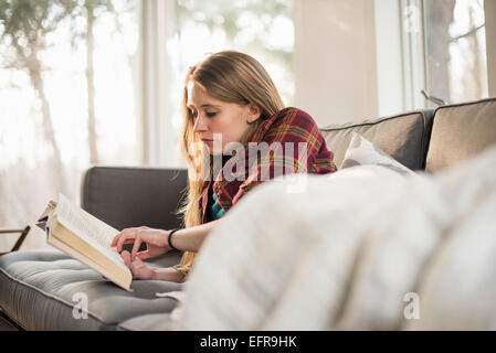 Giovane donna sdraiata su un divano, la lettura di un libro. Foto Stock