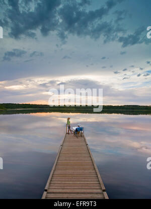 Un giovane uomo e donna seduta al termine di un lungo molo in legno raggiungendo fuori in un lago calmo, al tramonto. Foto Stock