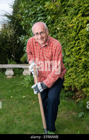 Uomo anziano in piedi in giardino, indossare abiti da lavoro e azienda vanga, felice Foto Stock