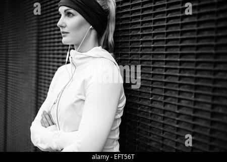 Immagine in bianco e nero di giovane donna che indossa gli auricolari inclinazione permanente di una parete che guarda lontano con fiducia. Femmina Fitness di riposo. Foto Stock