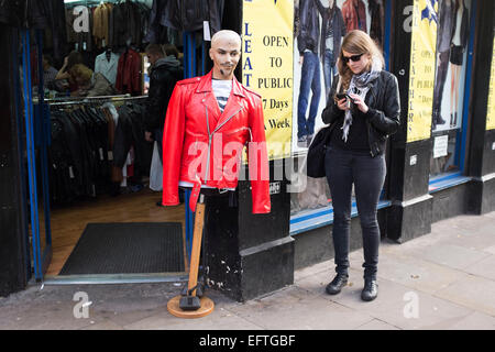 Ragazza texting mentre in piedi accanto ad un manichino che indossa un rosso giacca di pelle al di fuori di un negozio Il Brick Lane, Londra, Regno Unito. Foto Stock