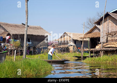 Intha uomo in proa presso il village con bambù tradizionali case su palafitte in Lago Inle, Nyaungshwe, Stato Shan, Myanmar / Birmania Foto Stock