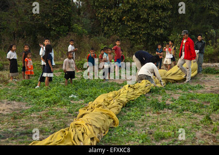 Vista orizzontale di giovani laotiani bambini aiutando a riporre una mongolfiera sbarcati vicino al loro villaggio. Foto Stock