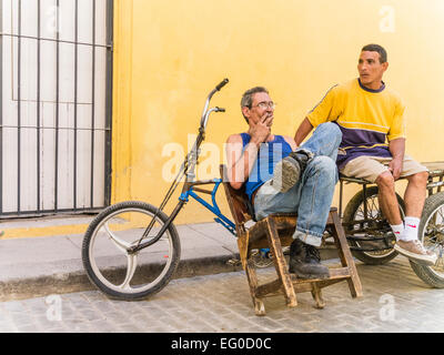 Due consegna biciclette uomini seduti a loro 3 ruote di biciclette di consegna contro una parete gialla e parlare a l Avana Vieja, Cuba. Foto Stock