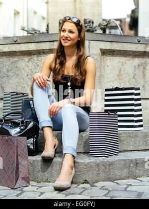 Foto di una giovane e bella donna seduta con il suo shopping bags presso una fontana in una vecchia città europea in appoggio. Foto Stock