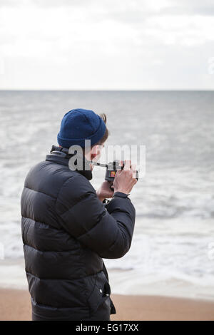 Giovane uomo che indossa 'puffa' giacca e cappello di lana su Chesil Beach in inverno scattare una fotografia con una fotocamera da 35mm. Foto Stock