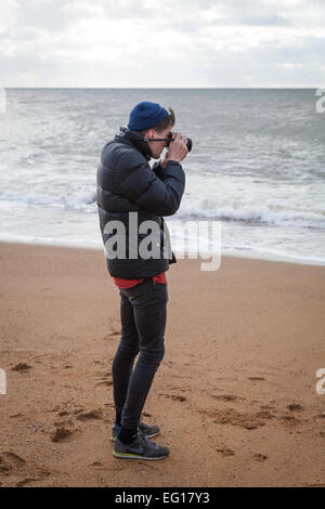 Giovane uomo che indossa 'puffa' giacca e cappello di lana su Chesil Beach in inverno scattare una fotografia con una fotocamera da 35mm. Foto Stock