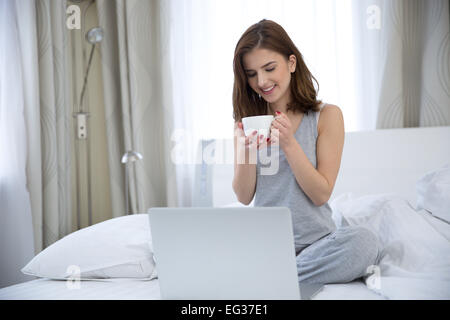 Ritratto di una donna sorridente con caffè e il computer portatile di seduta sul letto di casa Foto Stock