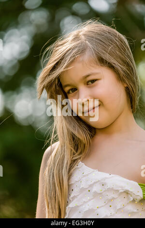 Ritratto di sette anni, con i capelli lunghi bambina indossa un abito senza maniche esterno, guardando molto coy e dolce Foto Stock