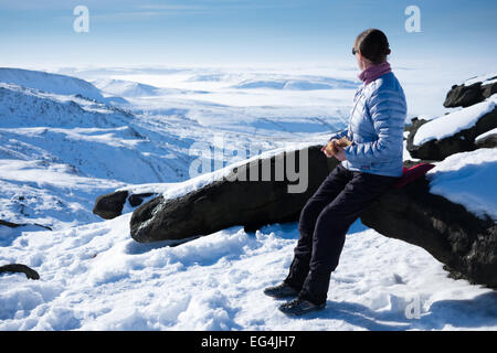 Una Donna vestita in outdoor gear si siede su una roccia nevoso guardando fuori attraverso un lansdscape di neve e cloud. Foto Stock