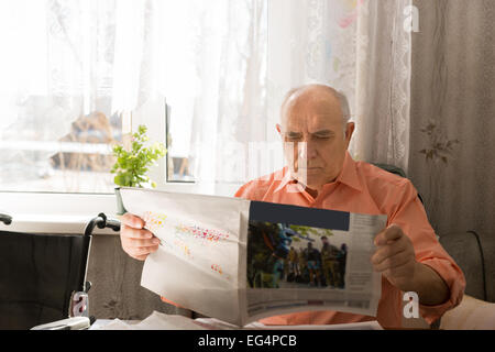 Close up seduta uomo Senior di lettura di articoli da Tabloid seriamente durante i momenti di relax presso il salotto vicino alla finestra con il bianco Foto Stock