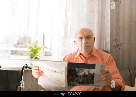 Close up grave uomo Senior Holding Tabloid guardando la telecamera mentre è seduto dentro la sua casa. Foto Stock