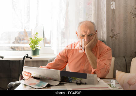 Close up focalizzata Vecchio seduto in soggiorno e durante la lettura del giornale con una mano sul suo volto. Foto Stock