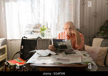 Grave uomo vecchio per leggere le notizie su Tabloid seriamente durante il riposo presso il salotto con la sua sedia a rotelle sul lato. Foto Stock