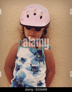 Felice piccola ragazza di capretto rosa in bici casco e occhiali da sole in piedi vicino al muro esterno Foto Stock