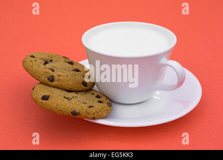 Due biscotti con scaglie di cioccolato su un piattino con latte intero in una tazza di caffè sulla cima di uno sfondo arancione Foto Stock