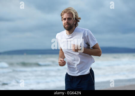 Un uomo nella sua 30's corre lungo una spiaggia con le nuvole scure sopra la testa Foto Stock