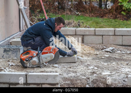 Un giovane maschio operaio operaio muratore misure un cinder breeze blocco breezeblock CMU con una sega circolare accanto a lui. Foto Stock