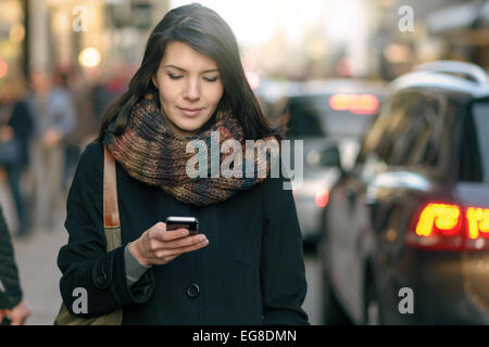 Moda giovane donna in nero cappotto e sciarpa colorata occupato con il suo telefono cellulare mentre si cammina per una via della città Foto Stock