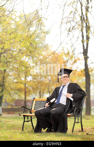 Colpo verticale di uno studente di college holding diploma e un libro seduti su un banco di lavoro in posizione di parcheggio Foto Stock