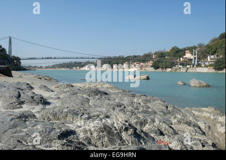 Un tratto roccioso di spiaggia a Rishikesh con Ram Jhula ponte in background che attraversa il Gange o Ganga river. Foto Stock