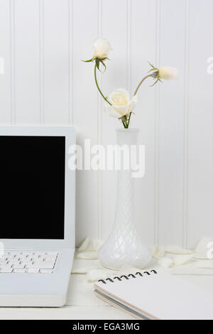 Primo piano di un bianco scrivania di legno con un computer portatile bianco e lattescente vaso con rose bianche. Una rilegatura a spirale di un pad alcuni petali di fiori sono n Foto Stock