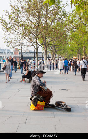 Uomo a suonare la chitarra sulla banca del sud con la folla che passa da Londra, Inghilterra Foto Stock