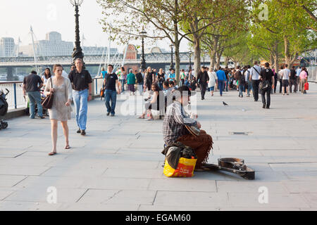 Uomo a suonare la chitarra sulla banca del sud con la folla che passa da Londra, Inghilterra Foto Stock