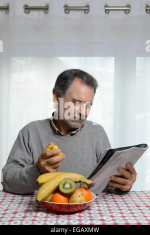 Uomo di mezza età leggendo una rivista mentre mangiando frutta per colazione in cucina a casa Foto Stock