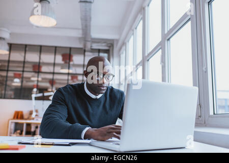 Inquadratura di un calvo imprenditore africano lavorando sul computer portatile in ufficio. Giovane web designer seduto alla sua scrivania di lavoro. Foto Stock