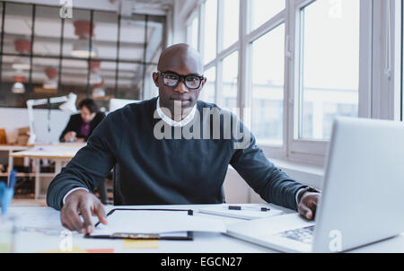 Ritratto di bello giovane africano alla sua scrivania con il computer portatile. L'uomo africano guardando la fotocamera mentre si è al lavoro. Foto Stock