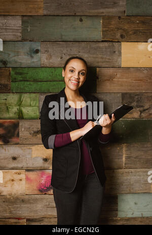 Ritratto di giovane attraente executive femmina con un blocco appunti in piedi contro una parete in legno in ufficio. Giovane americano africano