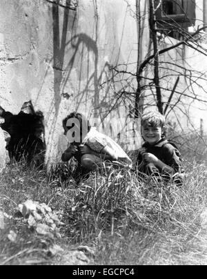 1930s 2 KIDS accanto al vecchio edificio ragazza spiata nel foro nella parete ragazzo seduto in erba sorridente Foto Stock