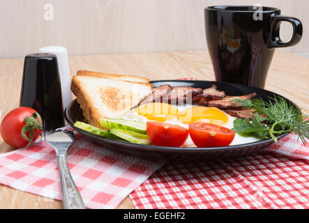 Uova fritte con pancetta, pane, caffè e le verdure sul piatto nero Foto Stock
